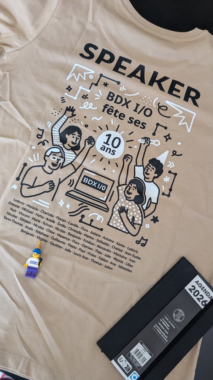 Photo d’un t-shirt “Speaker BDX I/O” comportant un dessin festif et les noms de tous les speakers de l’édition. Posés dessus, un lego représentant une speakeuse BDX I/O, et un petit agenda 2026.