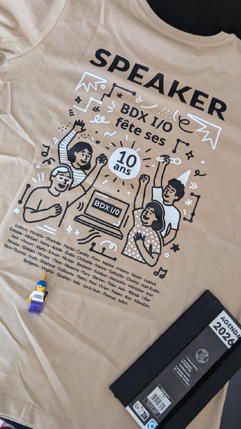 Photo d&rsquo;un t-shirt &ldquo;Speaker BDX I/O&rdquo; comportant un dessin festif et les noms de tous les speakers de l&rsquo;édition. Posés dessus, un lego représentant une speakeuse BDX I/O, et un petit agenda 2026.