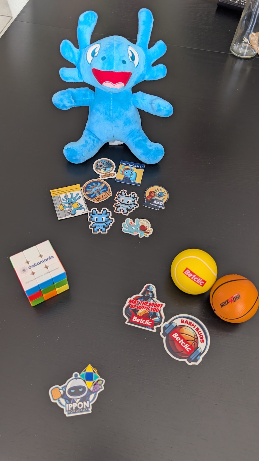 Photos de goodies posés sur une table : une peluche bleue en forme d’axolotl, un Rubik’s cube, des balles anti-stress et des stickers