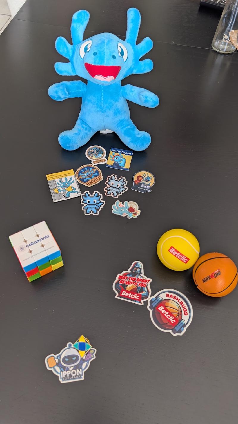 Photos de goodies posés sur une table : une peluche bleue en forme d&rsquo;axolotl, un Rubik&rsquo;s cube, des balles anti-stress et des stickers
